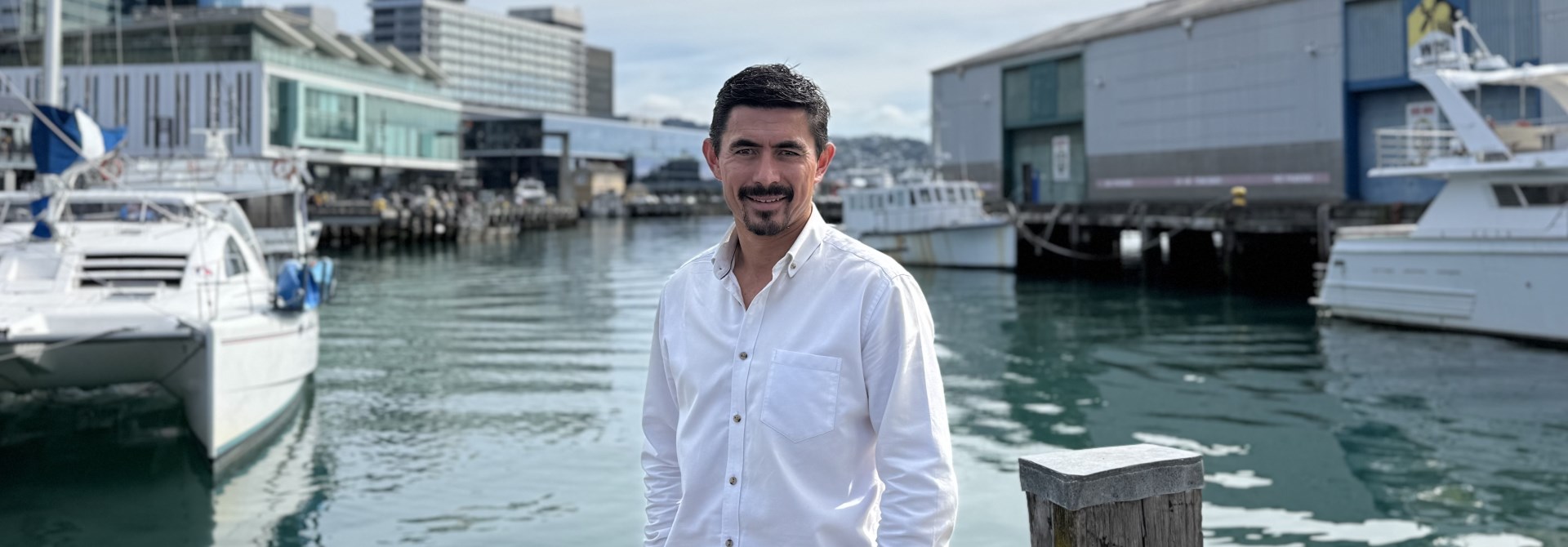 A man in a white shirt standing infront of the harbour with buildings in the background.