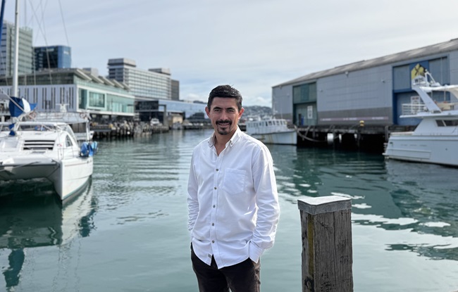 A man in a white shirt standing infront of the harbour with buildings in the background.