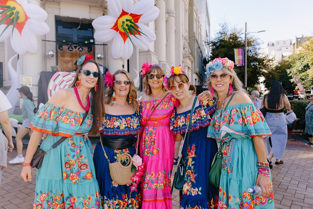 Five woman in colourful dresses infront a white building. 