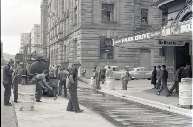Various stages of asphaltic road surfacing by Council workmen, Lambton Quay end of Featherston Street. People passing by have stopped to watch work in progress. Trolly buses in background