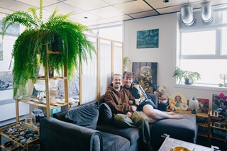 A couple sitting on a grey couch with a divder behind them and knick knacks on shelves around them.