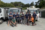 Group of men all standing together infront of trucks.