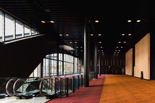 Inside of a building with orange and red carpet and escalators on the left hand side.