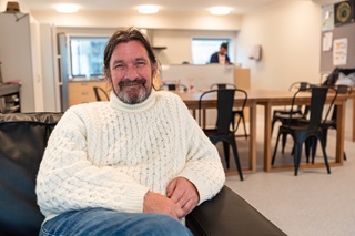 A smiling man with long hair and a beard wearing a white knitted sweater, sitting relaxed on a black couch in a communal living space with wooden tables and chairs in the background.