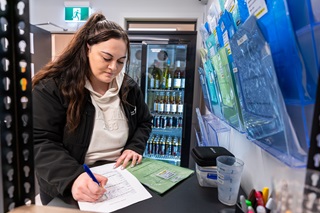 A young woman with long dark hair tied in a ponytail, wearing a black jacket and light hoodie, writing on a form at a counter beside a wall of document holders and a refrigerator filled with drinks.