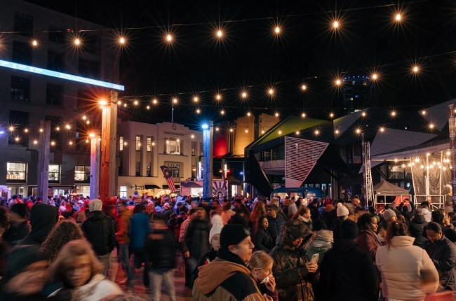 A crowd of people all dressed up in their warm winter clothing, moving around the waterfront at night.