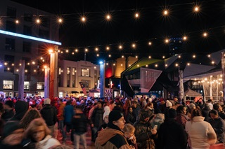 A crowd of people all dressed up in their warm winter clothing, moving around the waterfront at night.