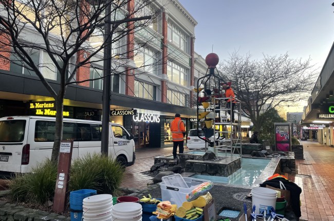 Buckets and cleaning equipment on the ground infront of a fountain where a person in high vis is cleaning.