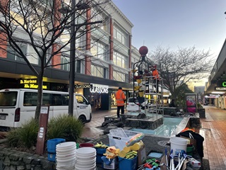 Buckets and cleaning equipment on the ground infront of a fountain where a person in high vis is cleaning.