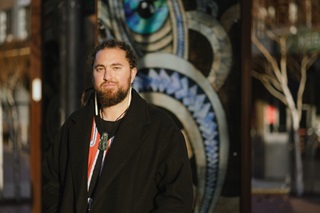 A person wearing a black jacket standing infront of their artwork in a light box exhibition.