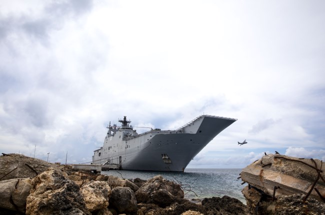 HMAS Canberra front at sea with rocks in foreground and plane in background.
