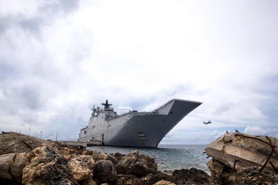 HMAS Canberra front at sea with rocks in foreground and plane in background.