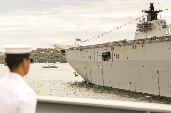 Ship sitting in the harbour with a person wearing a marine uniform looking at it.