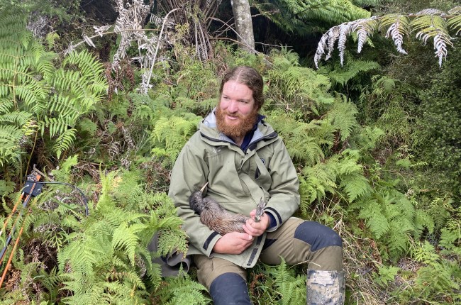 A man in a green jacket holding a kiwi in a bush.
