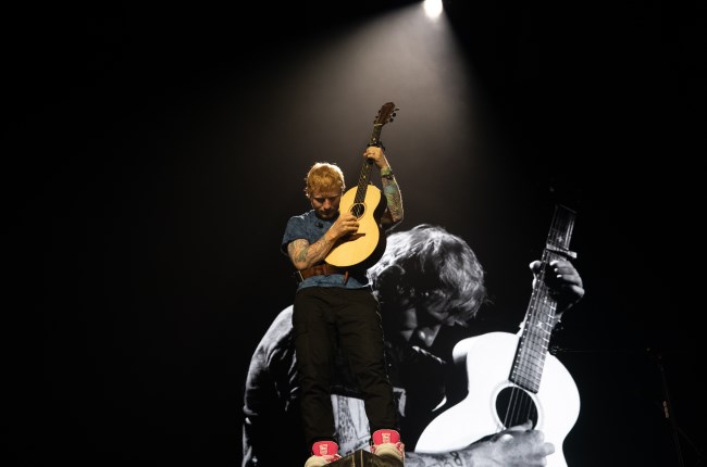 Ed Sheeran playing guitar on stage with projection of him in background.