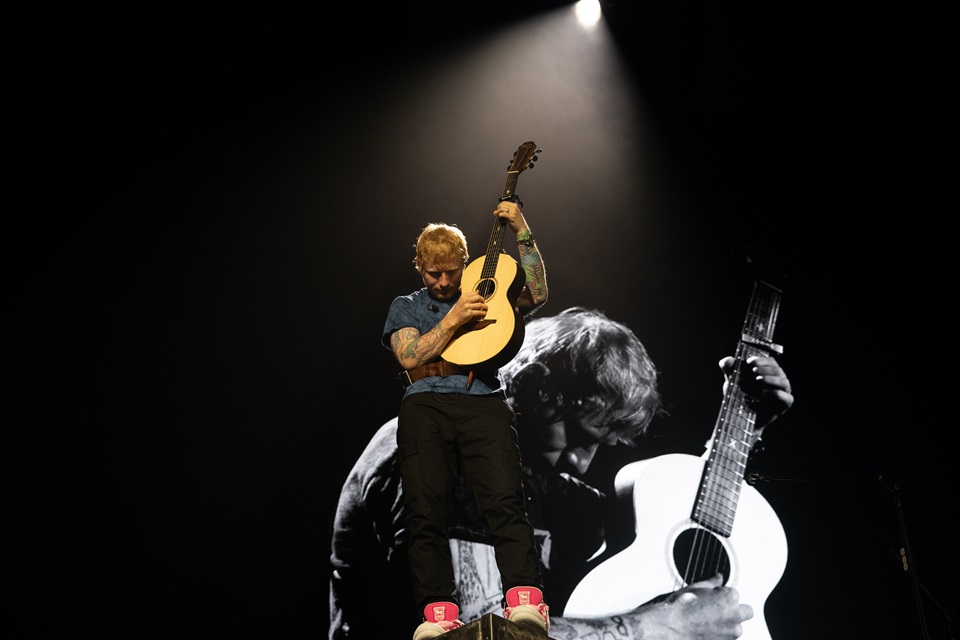 Ed Sheeran playing guitar on stage with projection of him in background.