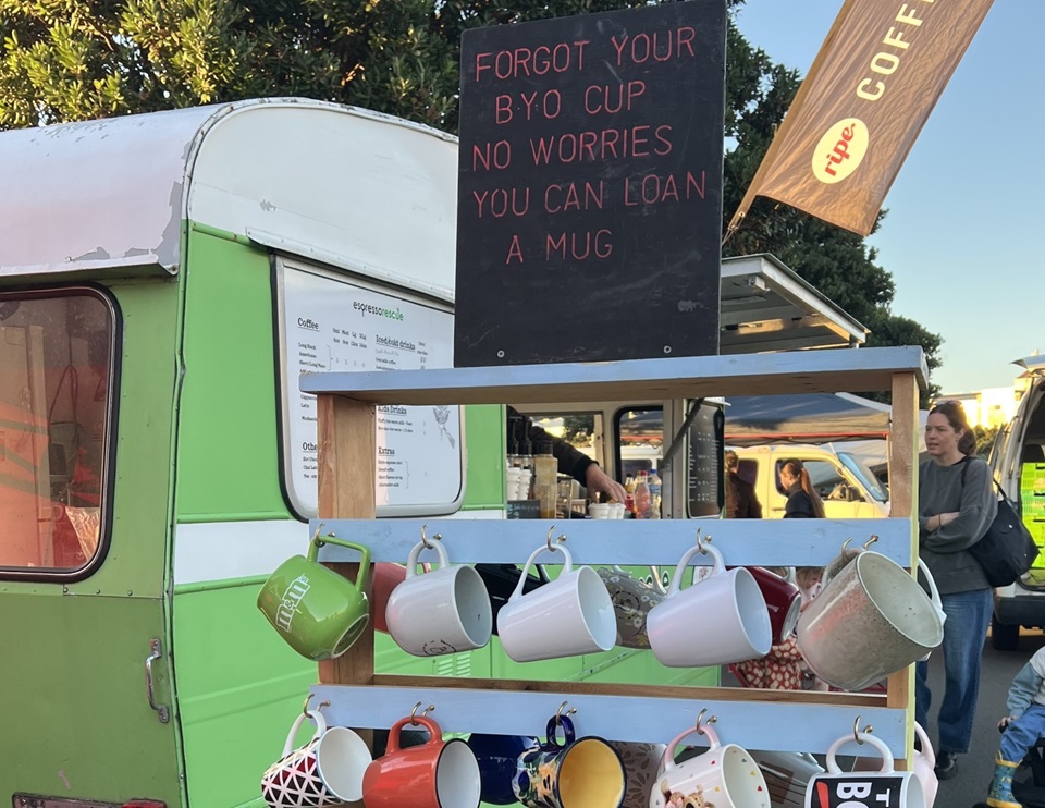 Mug.Cycle at Harbourside market with cups in forefront and signs and caravan in the background.