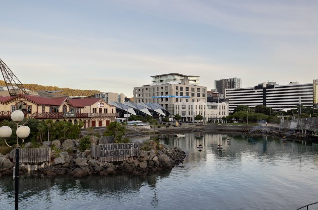 A grand two-storey wooden building, painted yellow with a red roof, beside large apartment buildings that overlook a reflective lagoon.