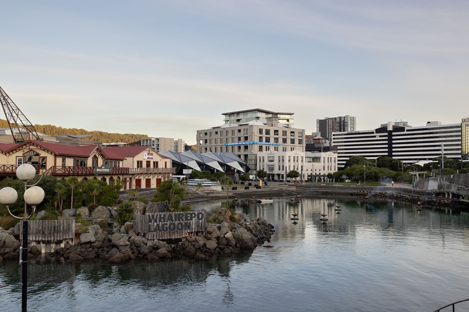 A grand two-storey wooden building, painted yellow with a red roof, beside large apartment buildings that overlook a reflective lagoon.
