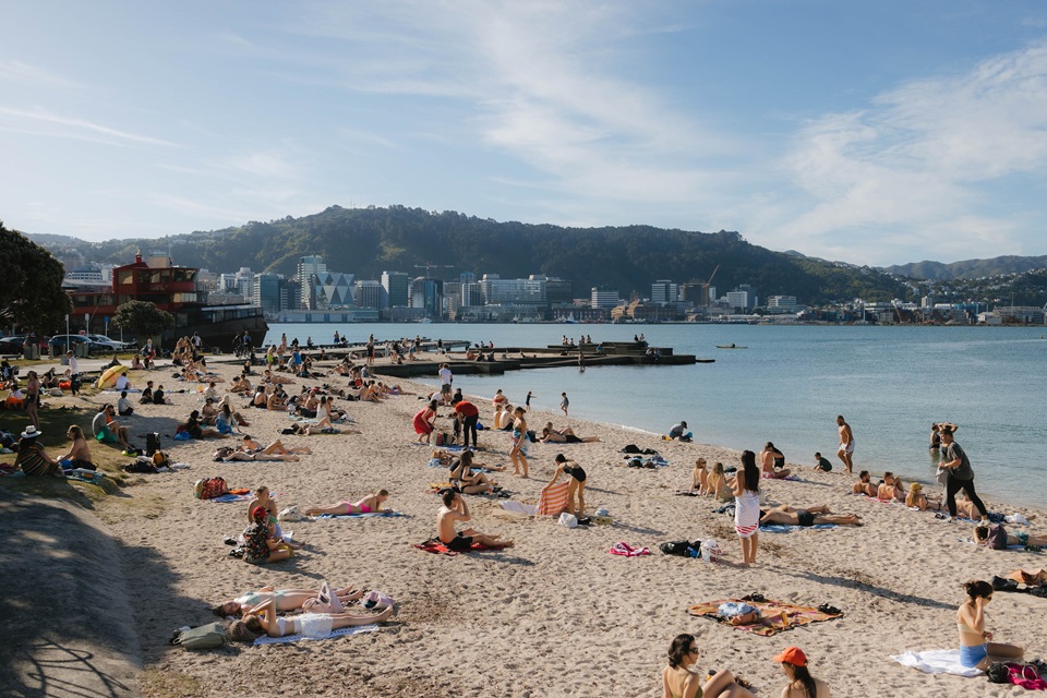 A crowded beach with golden sand on a sunny day, and across the harbour there are city buildings against tall hills.