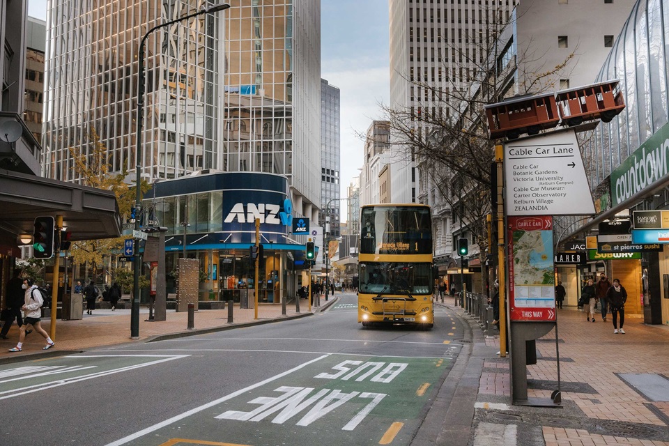 A yellow double decker bus drives along a designated bus lane through a paved street shadowed by tall city buildings on either side.