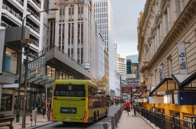 A green bus drives down a narrow road that is surrounded by tall city buildings and paved pedestrian paths on either side.