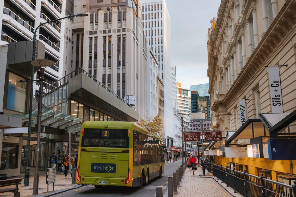 A green bus drives down a narrow road that is surrounded by tall city buildings and paved pedestrian paths on either side.