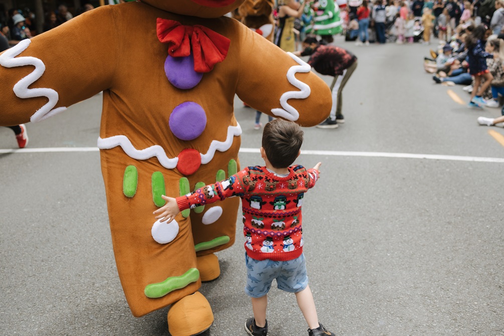 A performer dressed as a gingerbread man leans down to a young child running up to them with their arms open wide.