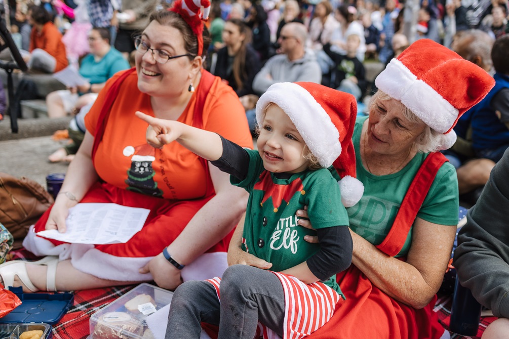 A family dressed in Christmas colours, wearing Santa hats enjoy a picnic while watching the performers.