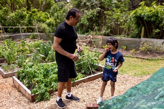 On the right, young boy enthusiastically eats a strawberry while a grandfather watches, smiling. They are standing among community garden growing plots at Innermost Gardens.