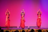 Three dancers in red dresses on stage with their hands raised above their heads.