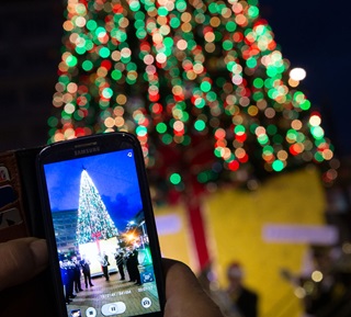 A Christmas tree at night time, all lit up with Christmas lights and someone standing in front of it taking a photo of it on their phone.