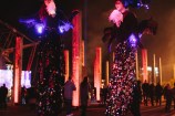 Two performers on stilts, adorned with lights and feathers.