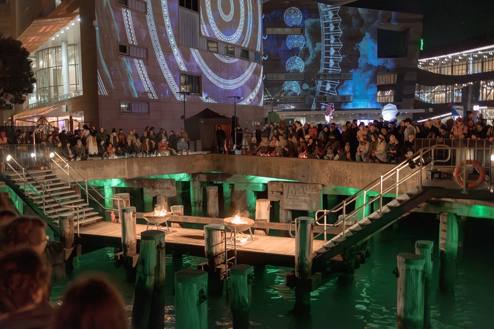 Two lit braziers on a platform bridge above the water with green mood lighting. A crowd is watching and Te Papa is lit in the background.