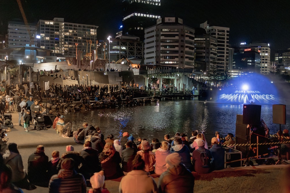 A large crowd of people seated around the edge of a lagoon watch movies projected on to a wall of water being sprayed from a fountain.