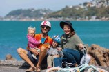 A family sit grinning on the side of Wellington harbour with helmets on and a kids bike lying in front of them.