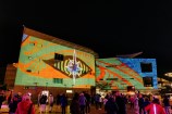 A crowd gather to watch a projection on the face of Te Papa. It is showing a giant stylised eye against a lime green background with orange/red accented Māori designs.