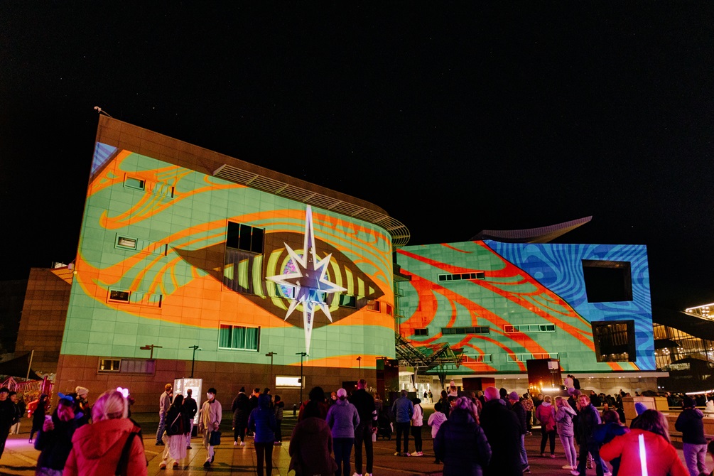 A crowd gather to watch a projection on the face of Te Papa. It is showing a giant stylised eye against a lime green background with orange/red accented Māori designs.