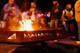 A large brazier made of weathered metal holds a roaring fire. The word 
