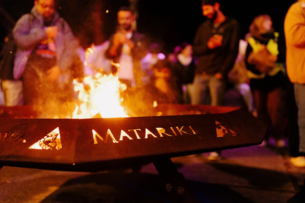 A large brazier made of weathered metal holds a roaring fire. The word 