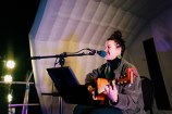 A woman performing on stage. She is strumming a guitar and singing into a microphone.