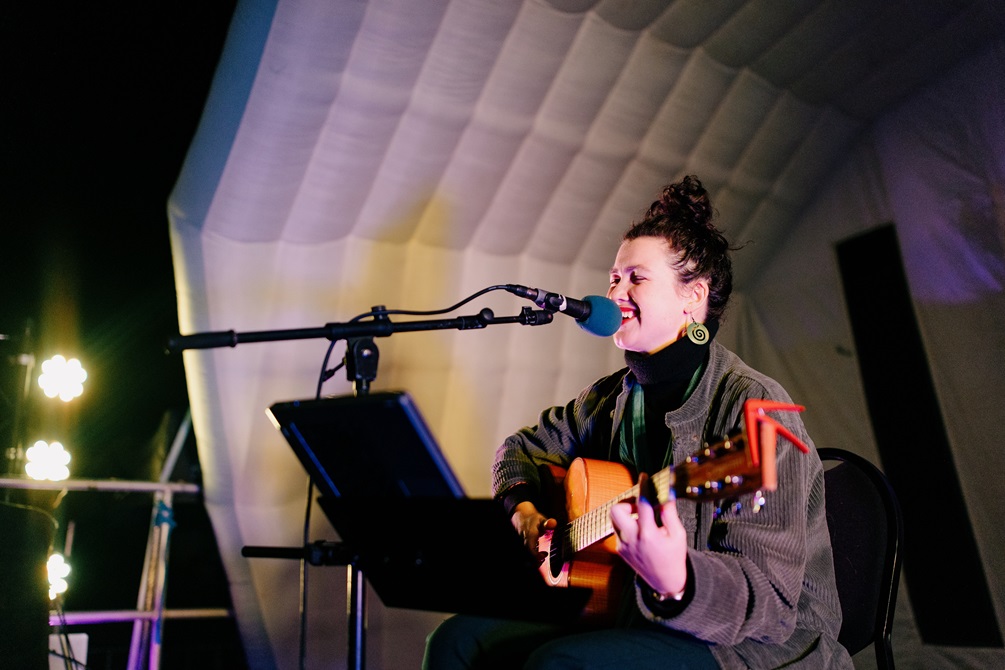 A woman performing on stage. She is strumming a guitar and singing into a microphone.