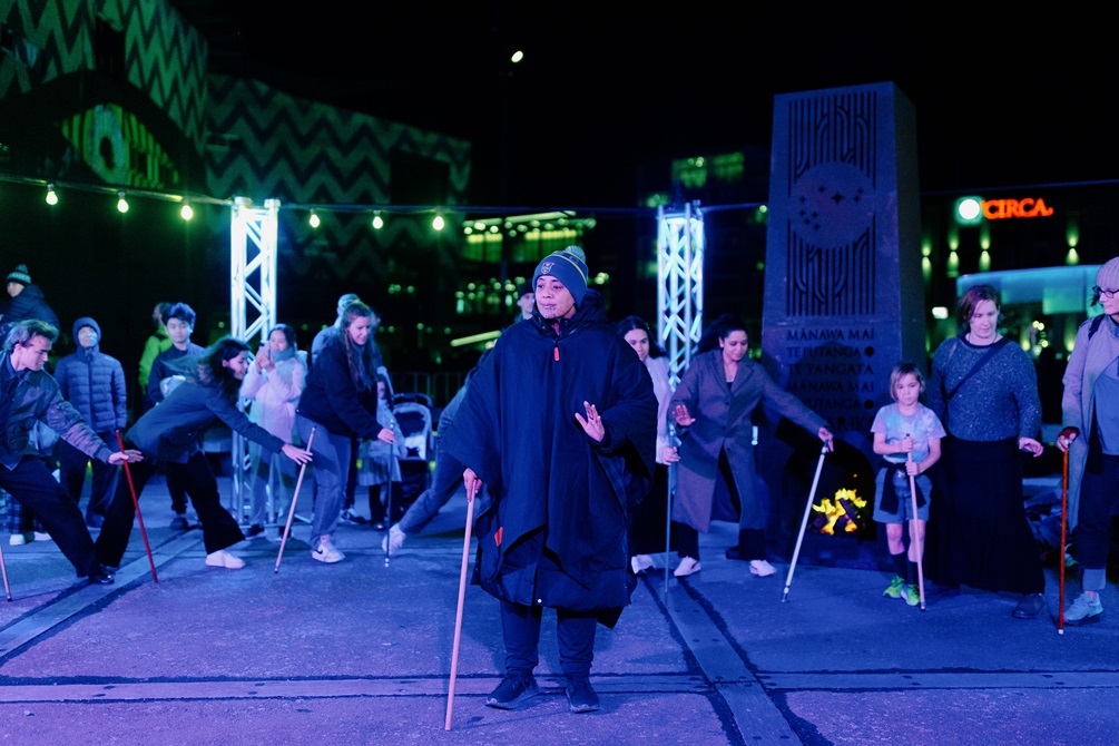 An instructor leading participants in a game involving balancing sticks. An unfortunate participant in the background is close to dropping theirs.