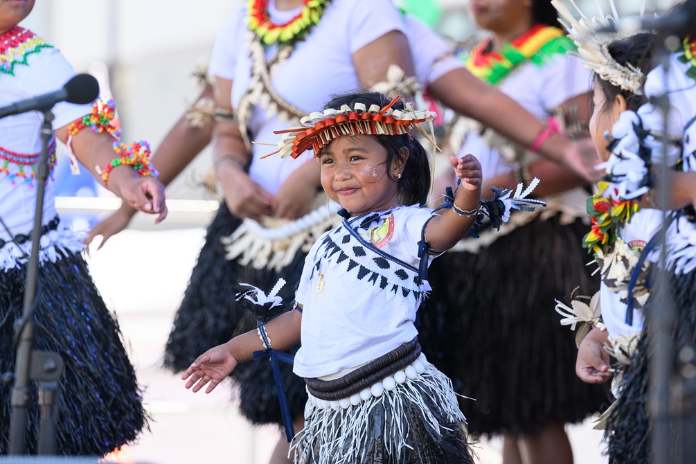 A child in traditional dress performs as part of a troupe with other performers in the background.