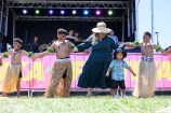 Children in grass skirts perform in front of the main stage. A woman and a small child in casual dress join them in the performance.