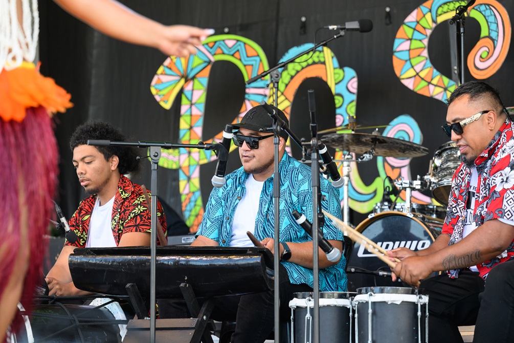 Three musicians dressed in colourful shirts play different types of drums on stage.
