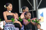 A group of girls in traditional dress perform on stage. They are wearing patterned skirts, black tops, and have garlands of green leaves on their wrists.