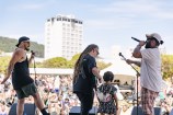 A band performs on stage with a large crowd on the grass in the background.