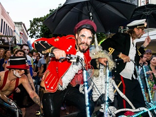 A two-man bike ridden by circus performers dressed in colourful attire and captains hats, surrounded by a large crowd on the middle of Cuba Street.