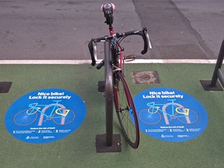 A bike is locked into a bike rack, and on the floor there are two signs with information about how to lock your bike securely.